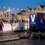 Mobilní kluziště na náměstí za mrazivého podvečera, bruslaři pod světly a stranou modrá elektrocentrála na přívěsu